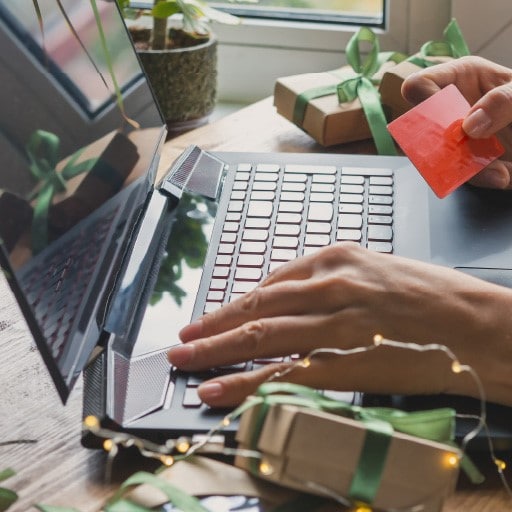 A person holiday shopping on a computer with their credit card and gifts all around